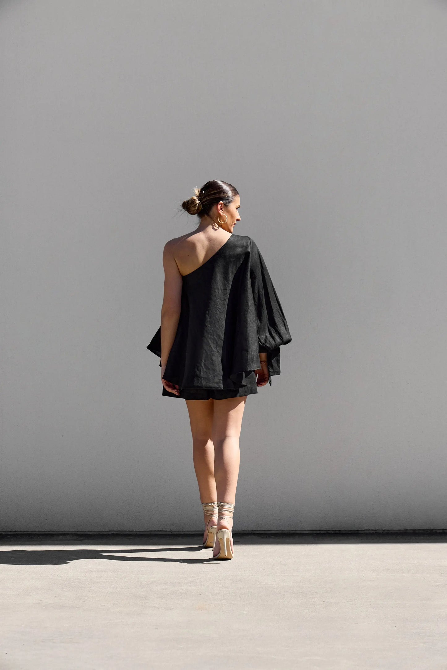 A woman stands outdoors against a white wall, wearing Isabella Longginous Black OS Balloon Sleeve Swing Top with an asymmetrical design. Her hair is in a bun, and she pairs the outfit with high-heeled sandals. Shadows dance under the sunlight.