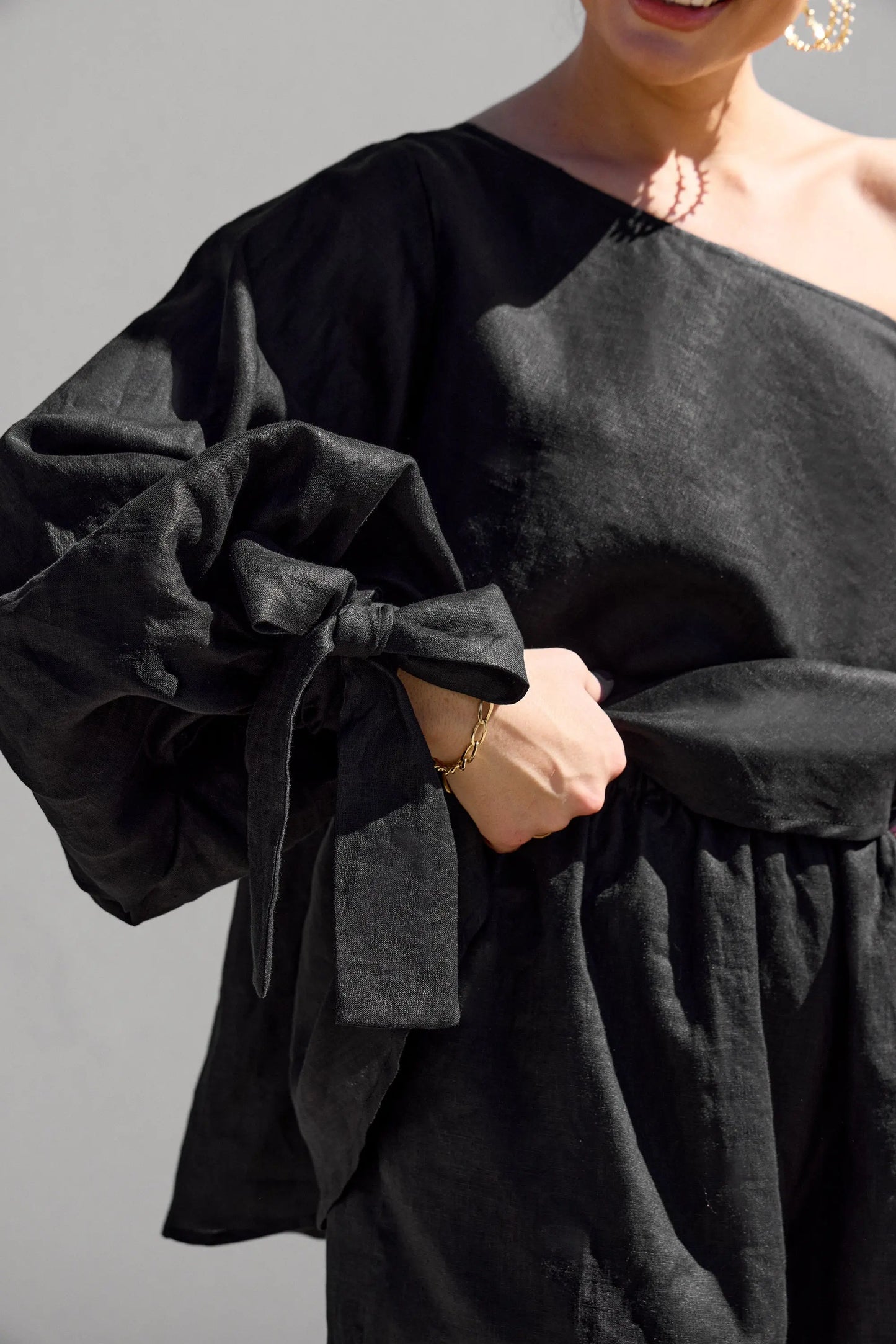 A person models the Isabella Longginou Black OS Balloon Sleeve Swing Top. The elegant off-the-shoulder design features a voluminous tied sleeve, held delicately at the waist against a plain light gray background.