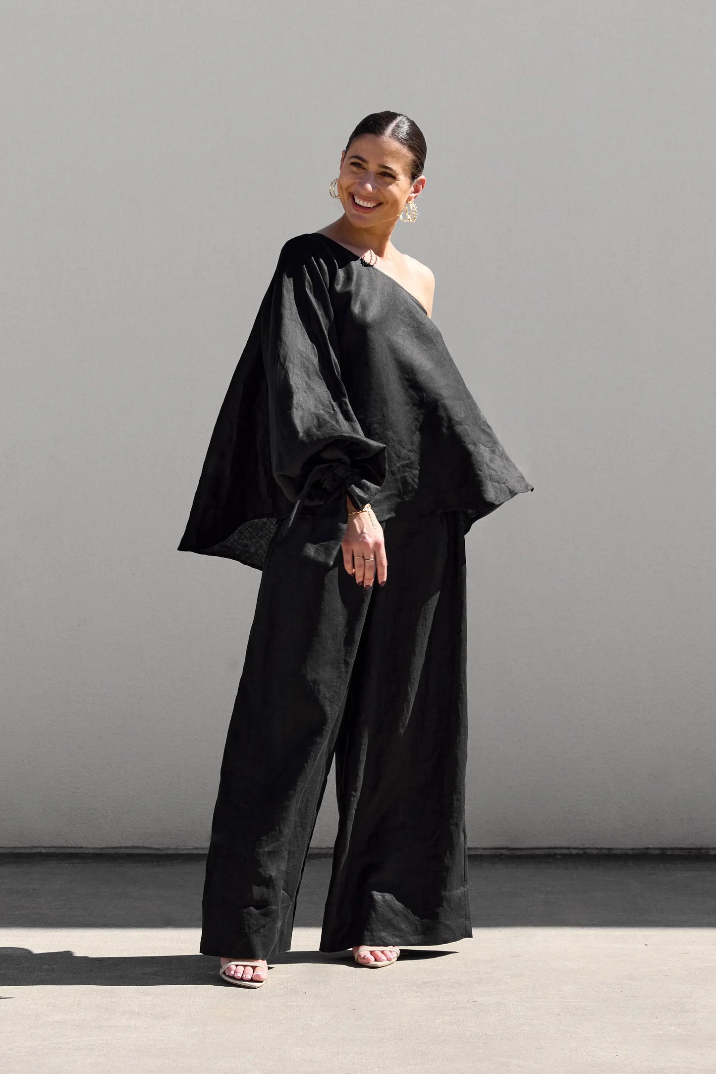 A person stands outdoors against a light wall, smiling. They wear an Isabella Longginou Black OS Balloon Sleeve Swing Top with wide-leg pants and high-heeled sandals. Their hair is pulled back, and they accessorize with large hoop earrings.