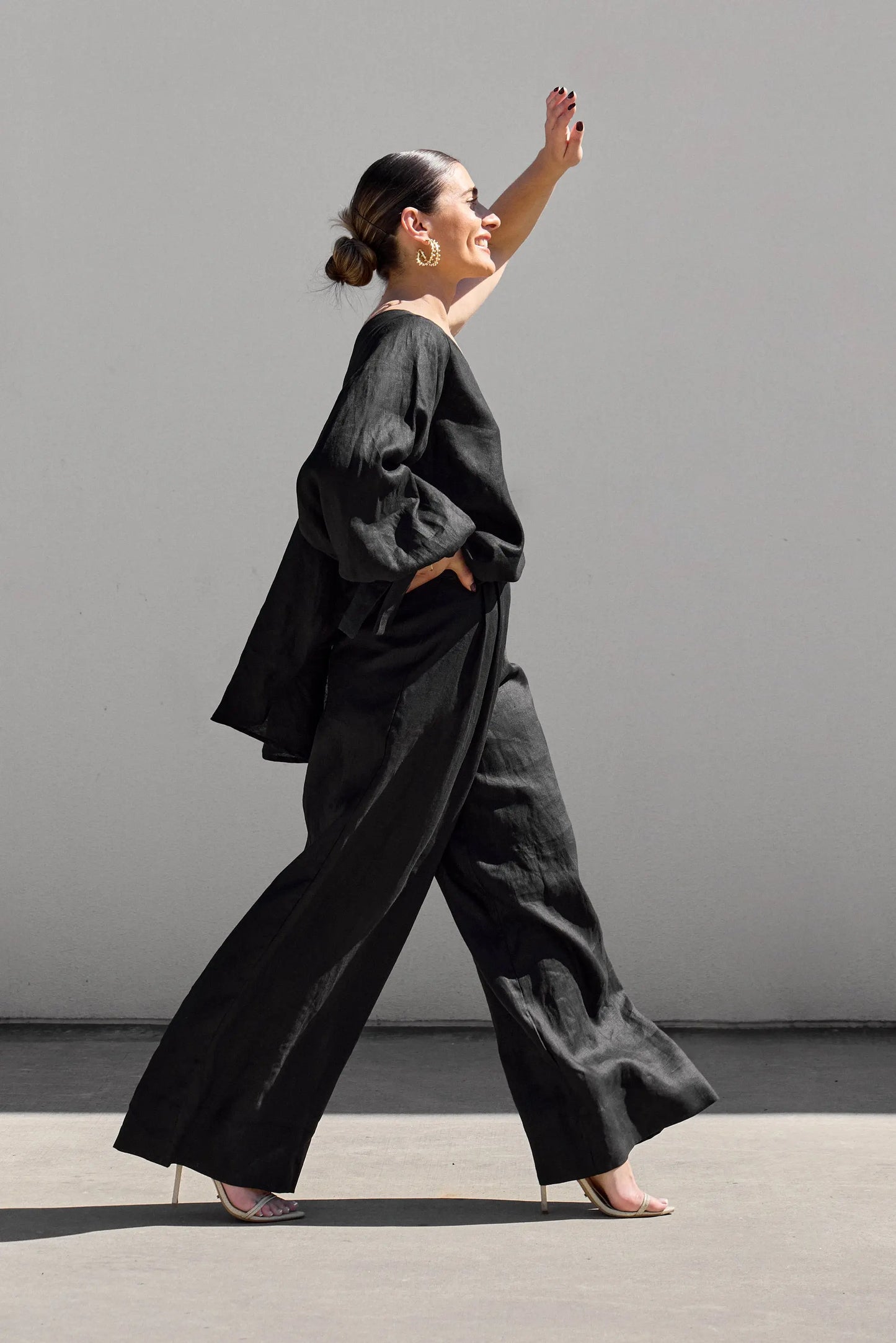 A woman confidently walks in profile against a gray backdrop wearing Black Palazzo Pants by Isabella Longginou, a loose top, and high heels. Her hair is styled in a bun as she raises her left arm with a smile.