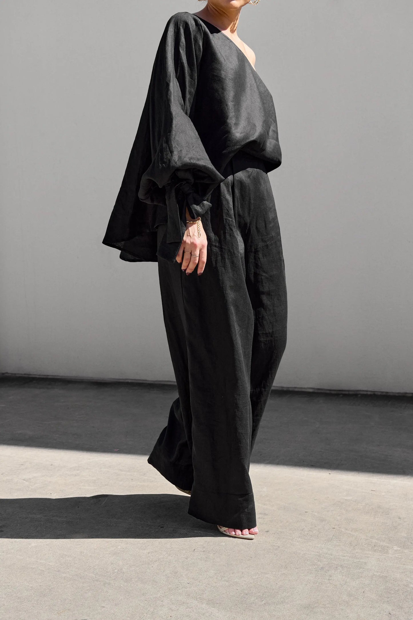 A person strolls on a concrete surface against a plain wall, showcasing Isabella Longginous one-shoulder black ensemble with loose-fitting long sleeves and stylish palazzo pants. Crafted from lightweight, flowy fabric, the outfit adds an elegant touch to the look.