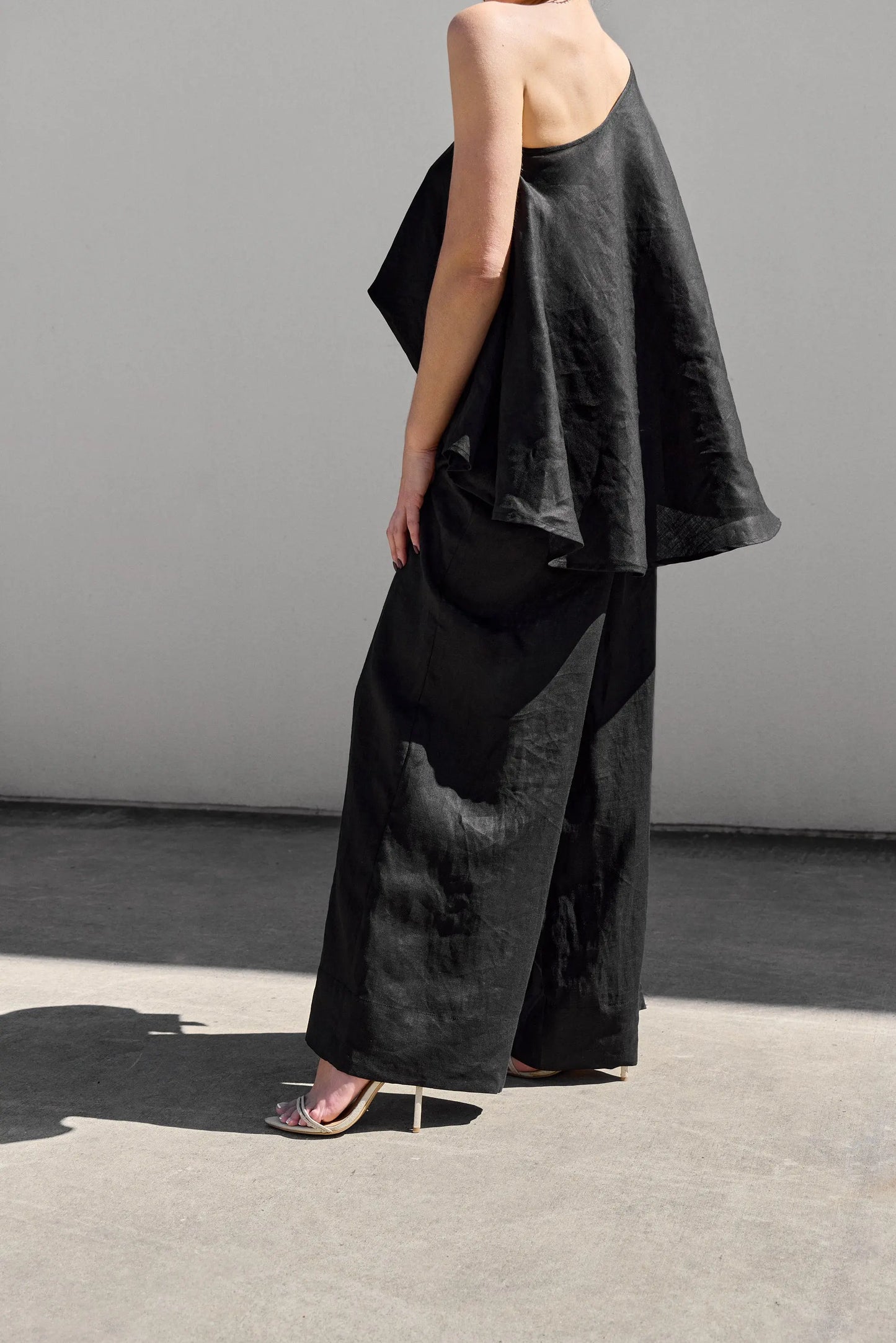 An individual stands on a sunlit concrete surface, donned in Isabella Longginous Black Palazzo Pants and an asymmetric black top with a flowing design. The outfit is completed with gleaming silver high-heeled sandals against a plain, light-colored wall.