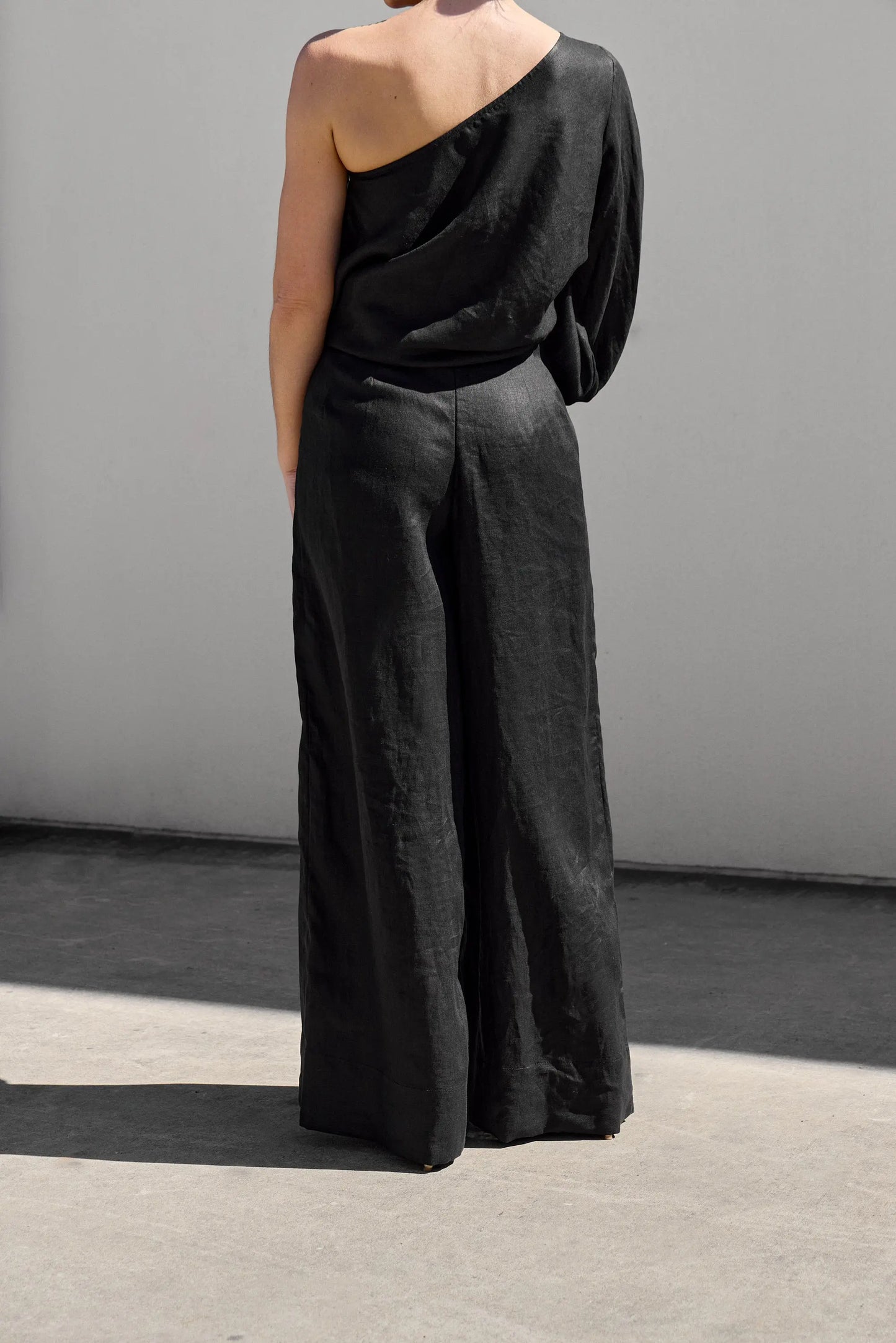 A person wearing Isabella Longginous Black Palazzo Pants and a one-shoulder black top stands with their back to the camera against a minimalist, light-colored wall. Shadows suggest sunlight.