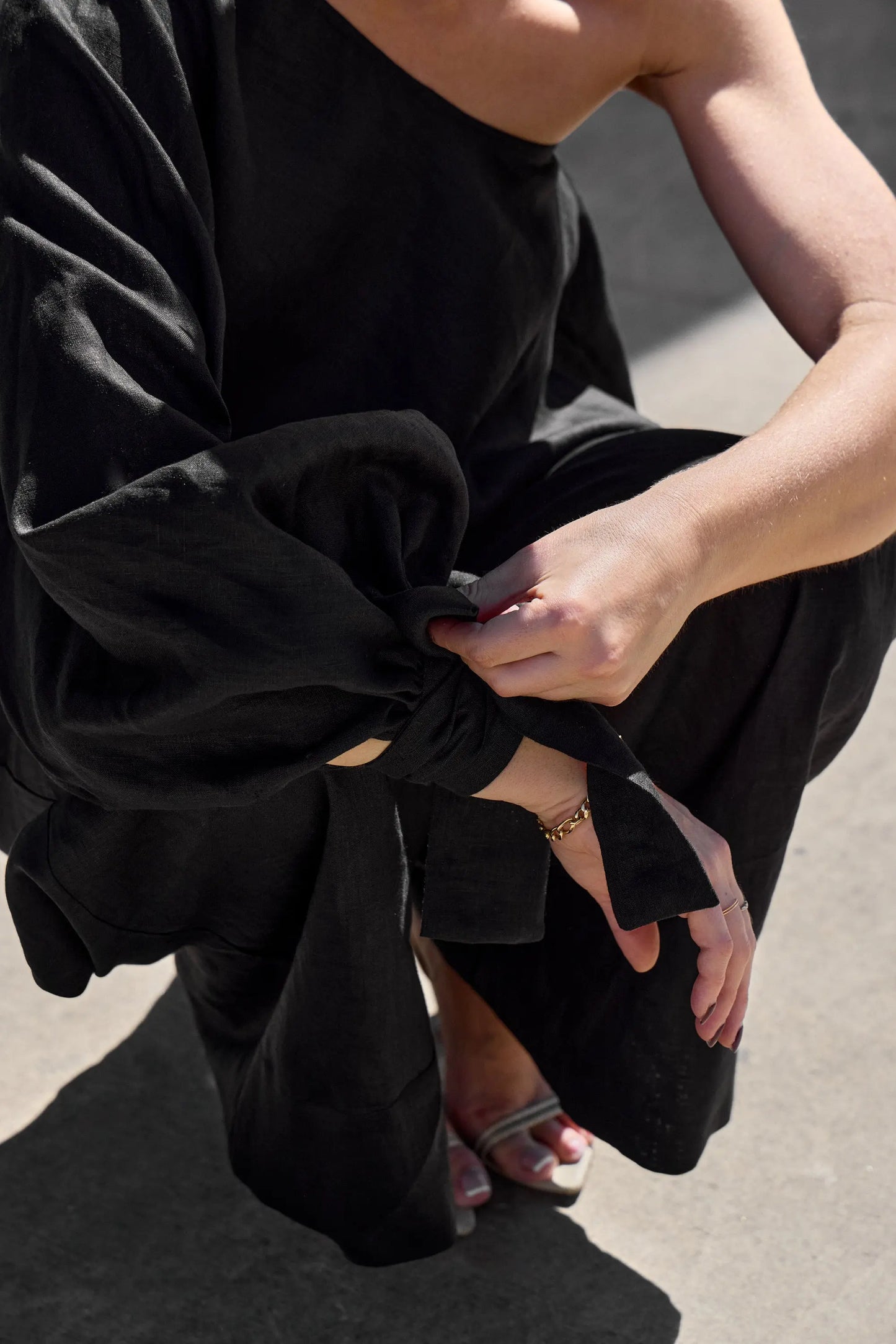 A person kneels on the concrete, adjusting the knot on an Isabella Longginou Black OS Balloon Sleeve Swing Top. They wear a gold bracelet on their right wrist and white open-toe sandals, with sunlight casting shadows around them.