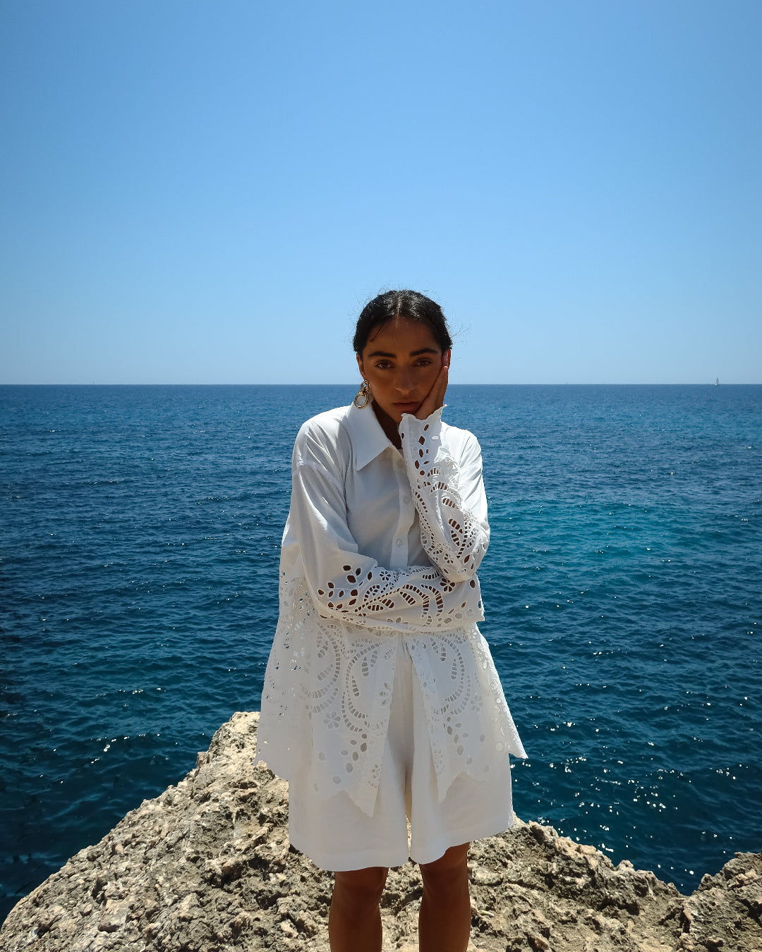 Person wearing a white dress with lace details standing on a rocky outcrop by the ocean.