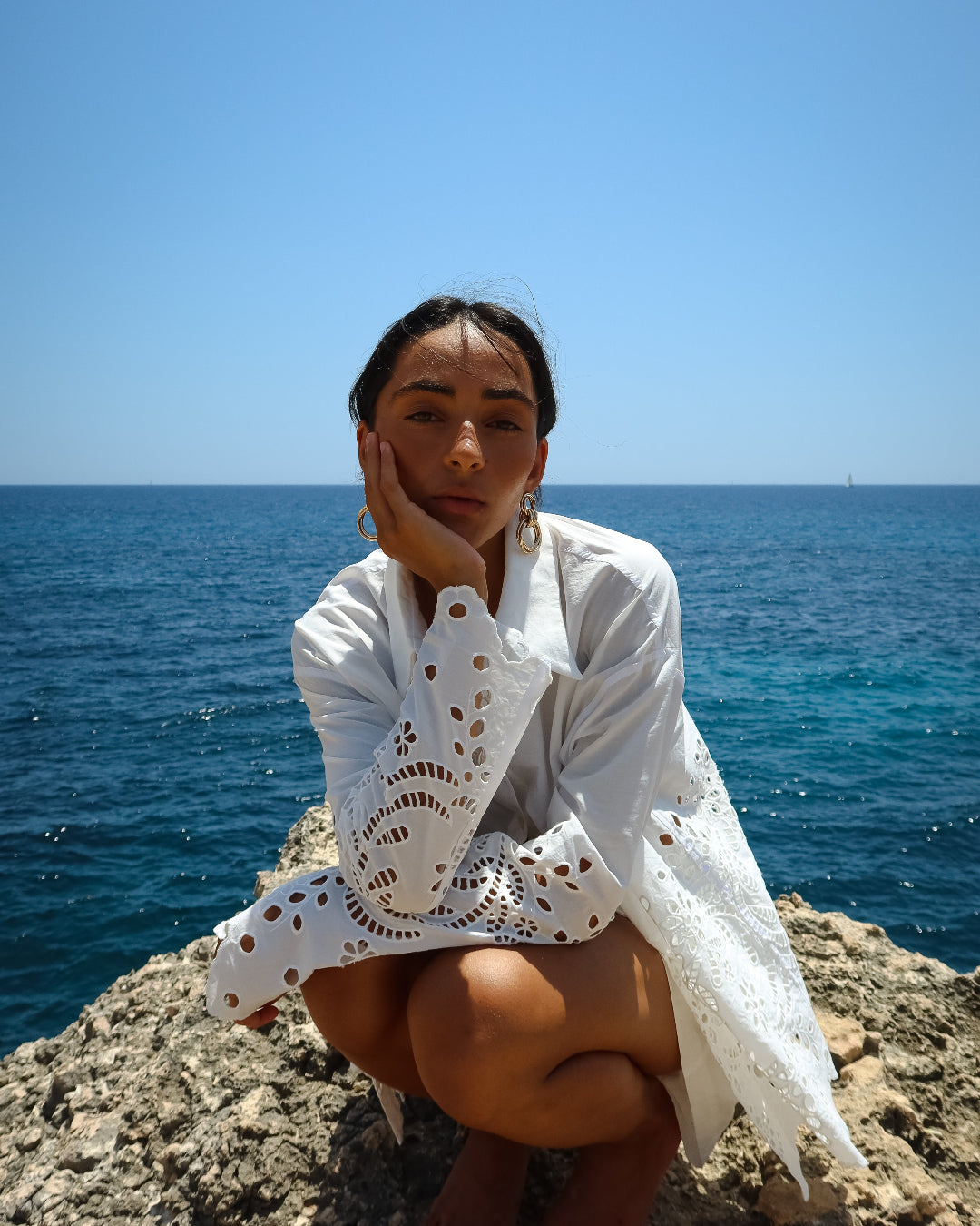 Woman in a white outfit sitting on a rocky outcrop with ocean and clear blue sky in the background