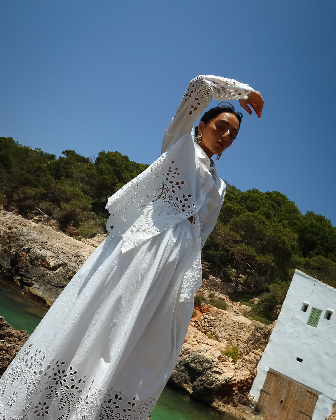 Woman in a white dress with lace details standing by a body of water with a clear blue sky.