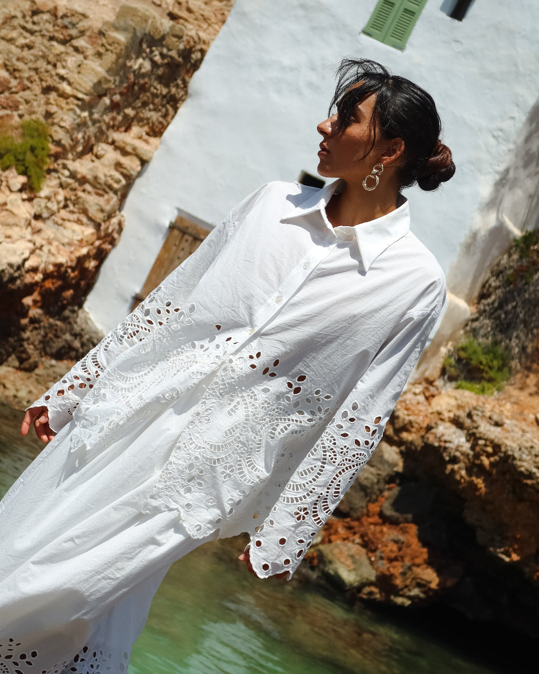 Woman wearing a white lace-trimmed shirt standing by a rocky coastline.