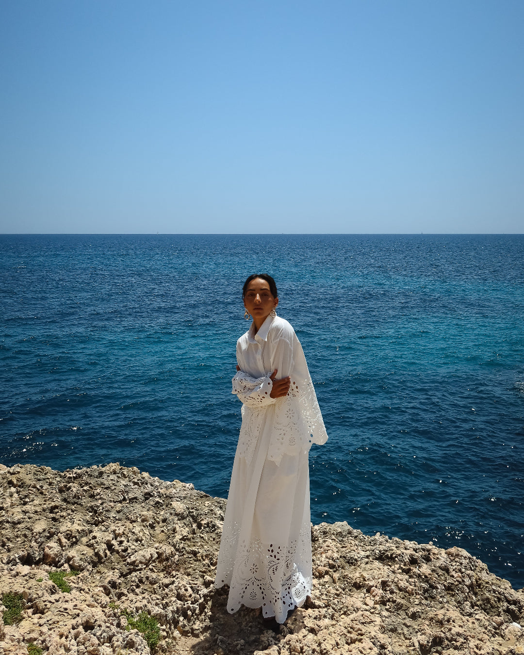 Person in a white dress standing on a rocky cliff overlooking the ocean.