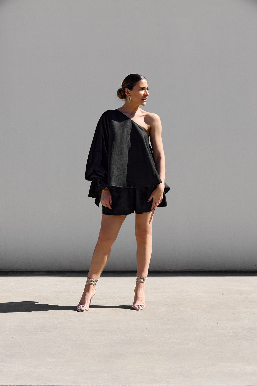 In front of a plain gray wall, a person with short hair wears Isabella Longginous Black OS Balloon Sleeve Swing Top paired with sandals. Facing slightly to the side in a relaxed pose, the sunny outdoor setting highlights their effortlessly chic style.