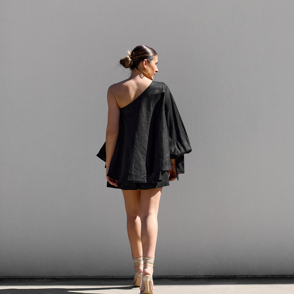 
                      
                        A woman stands outdoors against a white wall, wearing Isabella Longginous Black OS Balloon Sleeve Swing Top with an asymmetrical design. Her hair is in a bun, and she pairs the outfit with high-heeled sandals. Shadows dance under the sunlight.
                      
                    