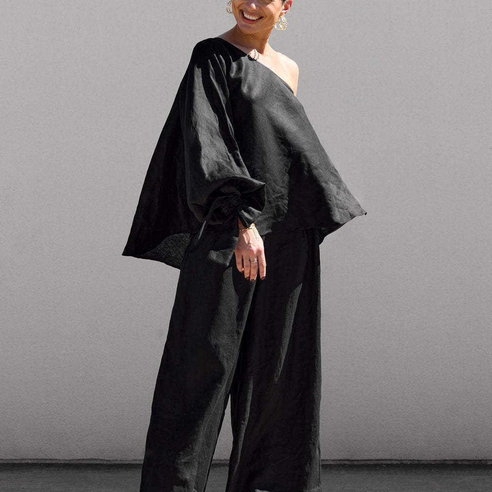 
                      
                        A person stands outdoors against a light wall, smiling. They wear an Isabella Longginou Black OS Balloon Sleeve Swing Top with wide-leg pants and high-heeled sandals. Their hair is pulled back, and they accessorize with large hoop earrings.
                      
                    