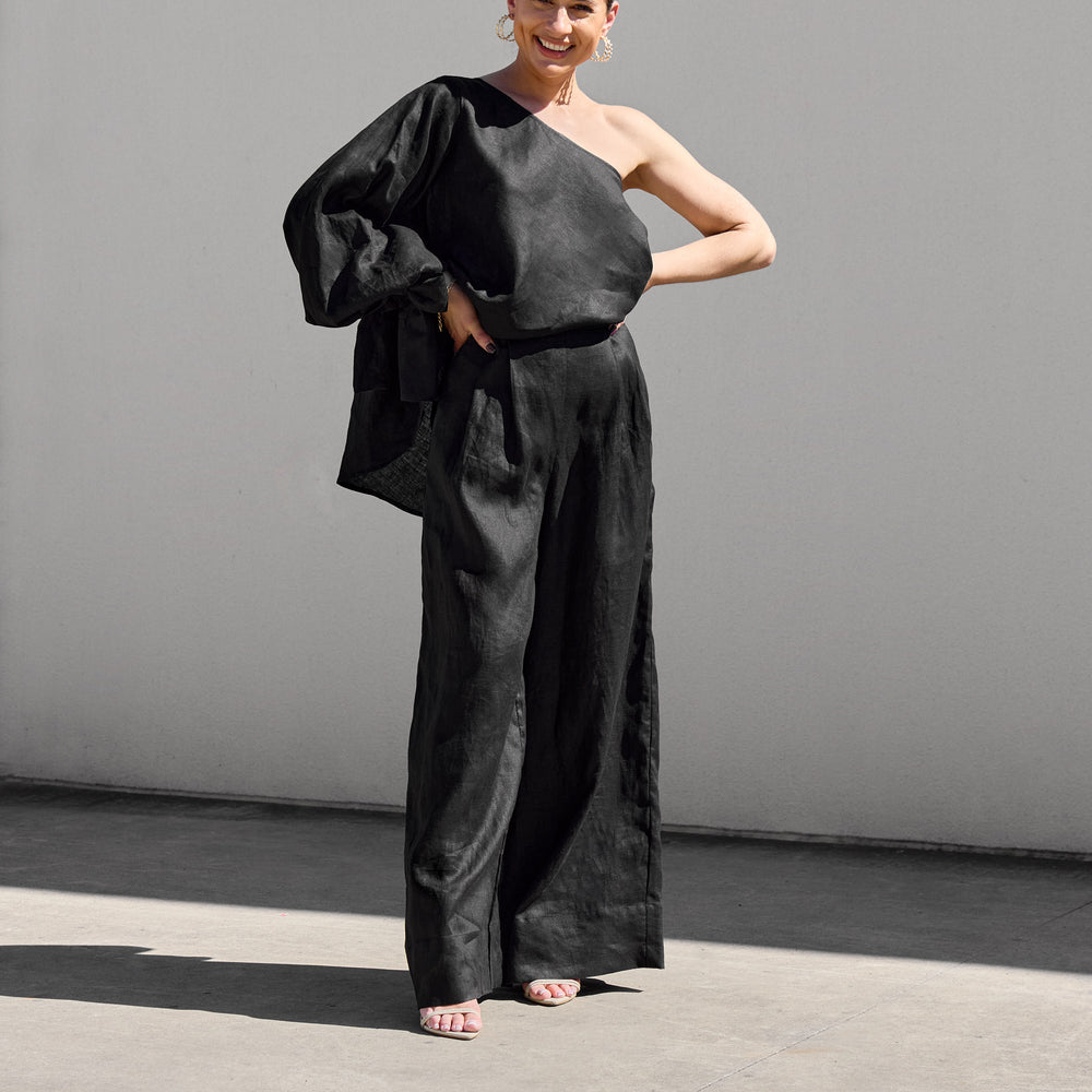 
                      
                        A woman exudes confidence outdoors in Isabella Longginou Black Palazzo Pants and a one-shoulder black top. Her hair is tied back, and she smiles with hands on hips against a light gray wall.
                      
                    