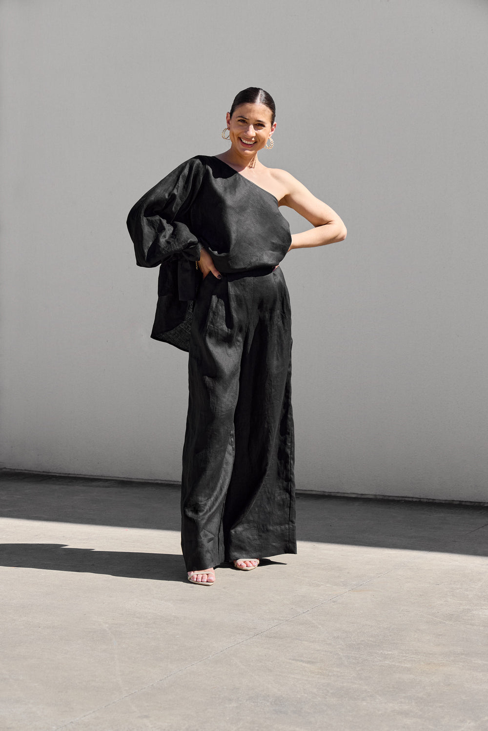 A woman exudes confidence outdoors in Isabella Longginou Black Palazzo Pants and a one-shoulder black top. Her hair is tied back, and she smiles with hands on hips against a light gray wall.