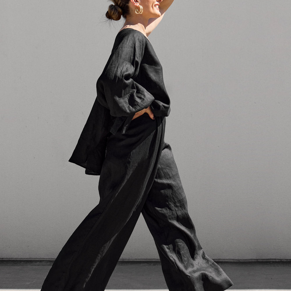 
                      
                        A woman confidently walks in profile against a gray backdrop wearing Black Palazzo Pants by Isabella Longginou, a loose top, and high heels. Her hair is styled in a bun as she raises her left arm with a smile.
                      
                    