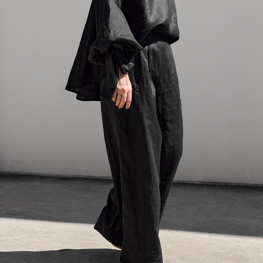 
                      
                        A person strolls on a concrete surface against a plain wall, showcasing Isabella Longginous one-shoulder black ensemble with loose-fitting long sleeves and stylish palazzo pants. Crafted from lightweight, flowy fabric, the outfit adds an elegant touch to the look.
                      
                    