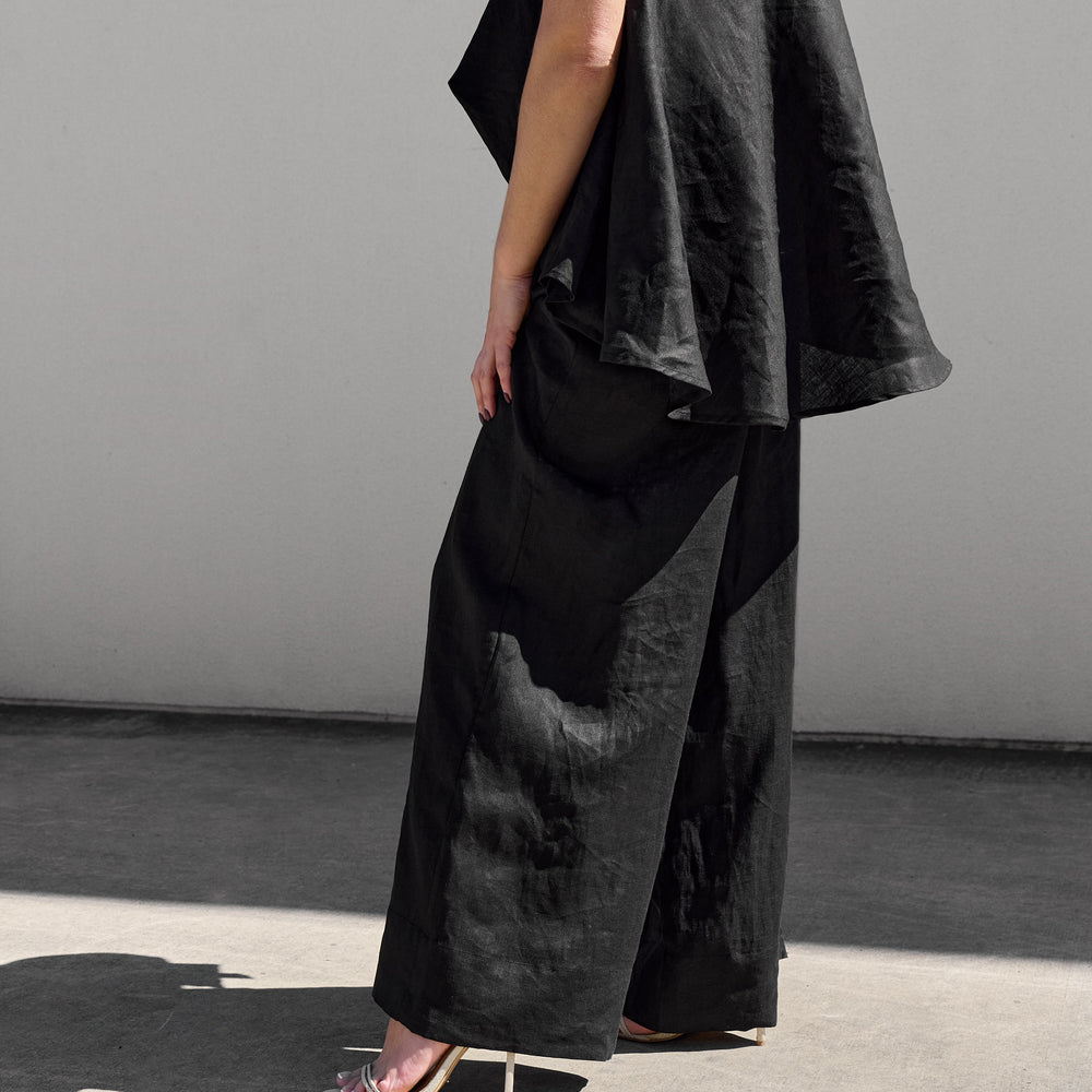 
                      
                        An individual stands on a sunlit concrete surface, donned in Isabella Longginous Black Palazzo Pants and an asymmetric black top with a flowing design. The outfit is completed with gleaming silver high-heeled sandals against a plain, light-colored wall.
                      
                    