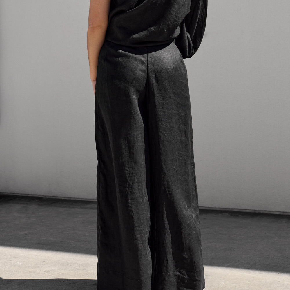 
                      
                        A person wearing Isabella Longginous Black Palazzo Pants and a one-shoulder black top stands with their back to the camera against a minimalist, light-colored wall. Shadows suggest sunlight.
                      
                    